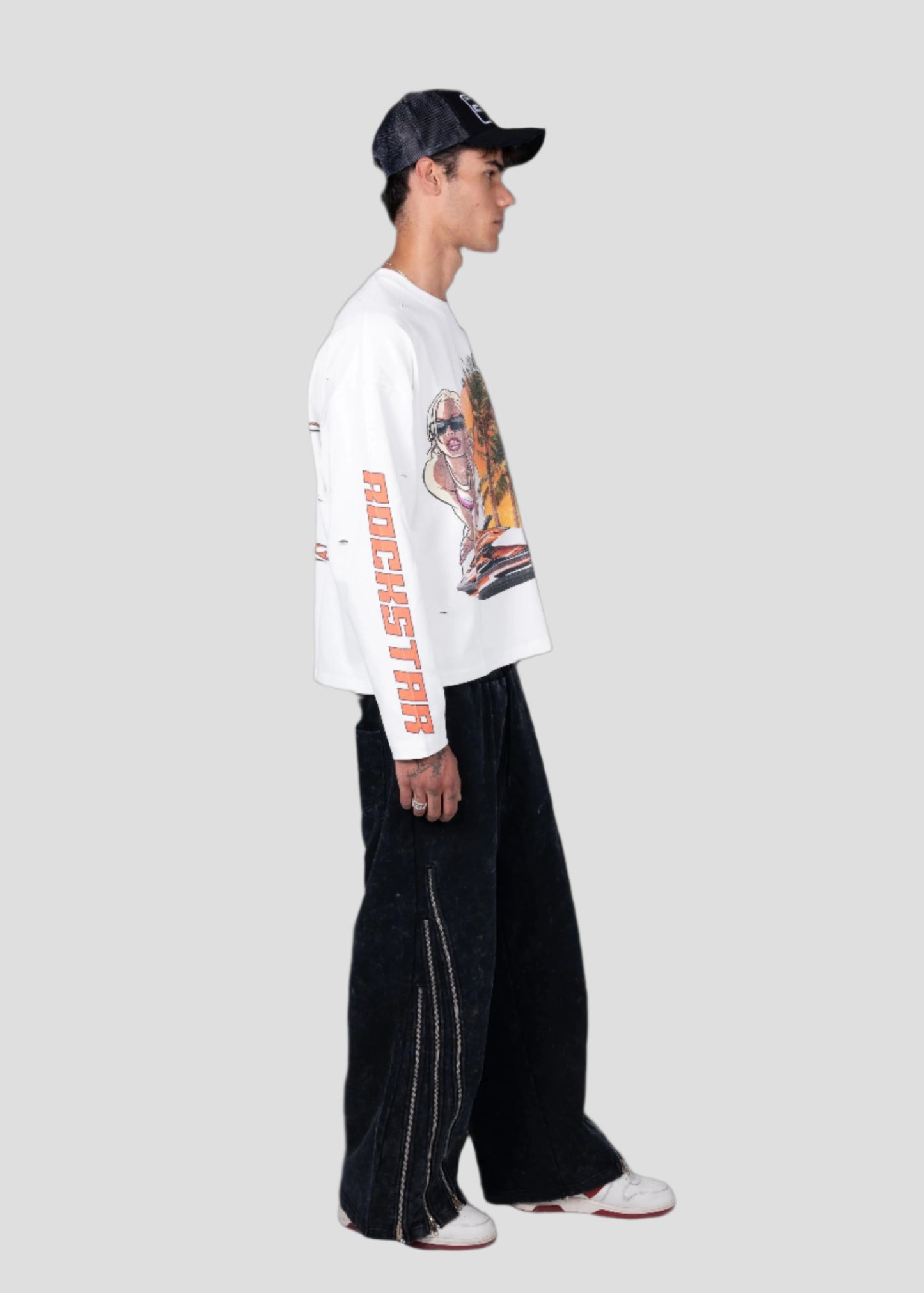 Person wearing a white long-sleeve shirt with graphics and text, black pants, and a cap on a black background
